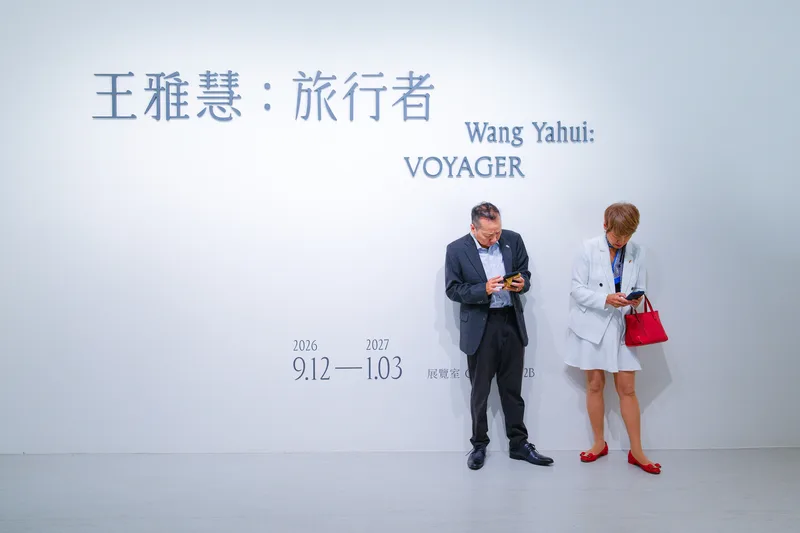 Two visitors absorbed in their phones beneath the title wall of an exhibition called Voyager